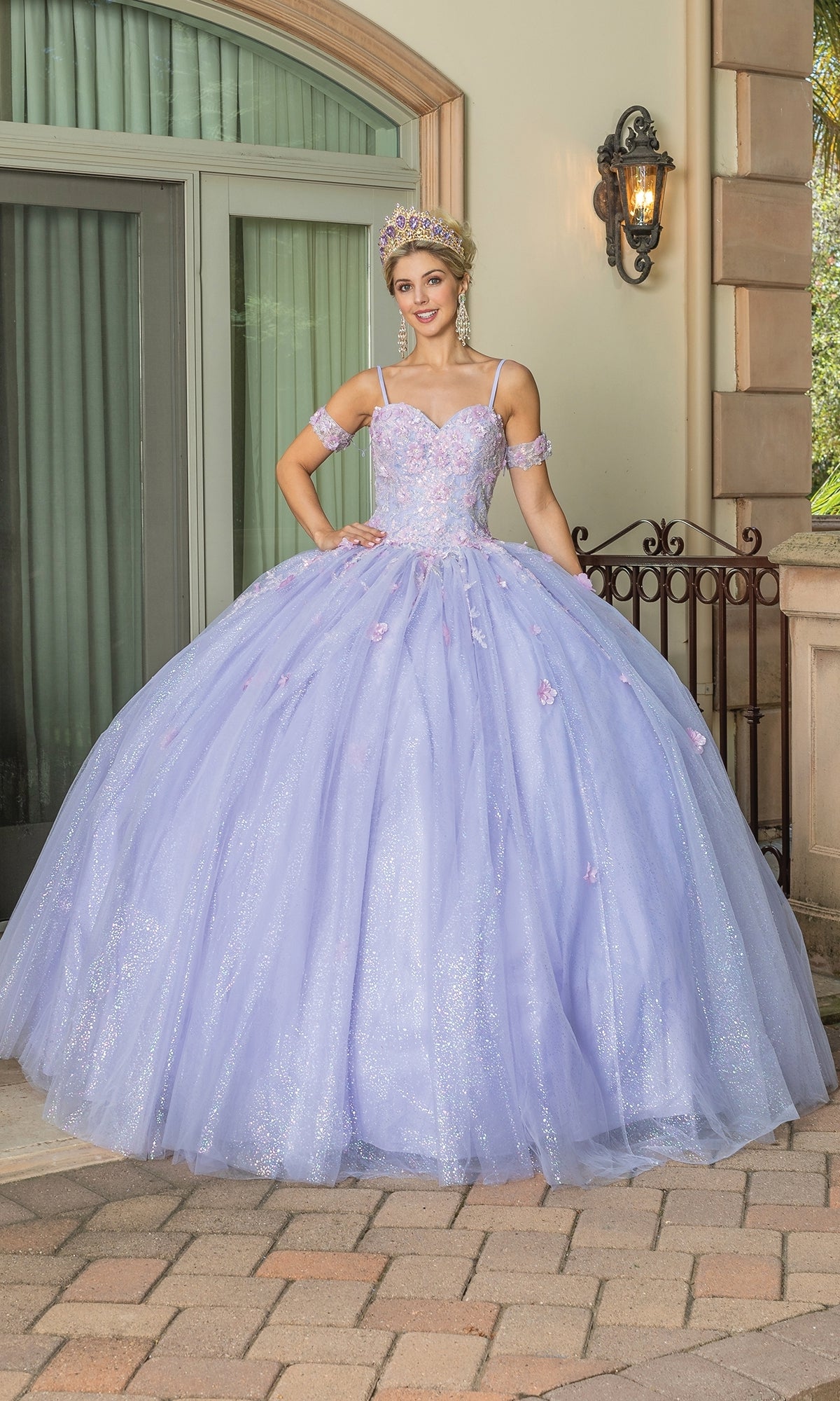 A young woman in the Quinceanera Dress 1785 By Dancing Queen, featuring a glittery lilac ball gown and sweetheart neckline, with matching arm cuffs and a tiara, stands outside near stone columns and a wrought iron railing.
